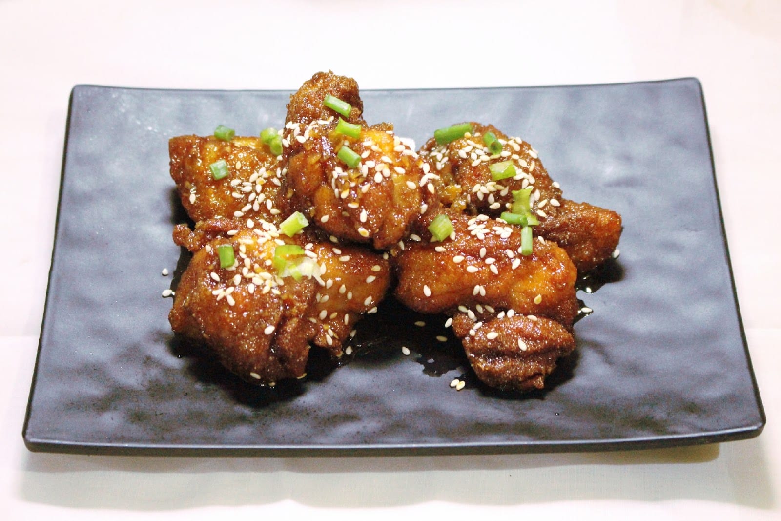 Fried chicken pieces glazed in sauce and topped with sesame seeds and spring onion, on a black plate